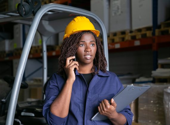 Focused Black warehouse worker in yellow hardhat standing near forklift and talking on cell. Shelves with goods in background. Medium shot. Labor or communication concept
